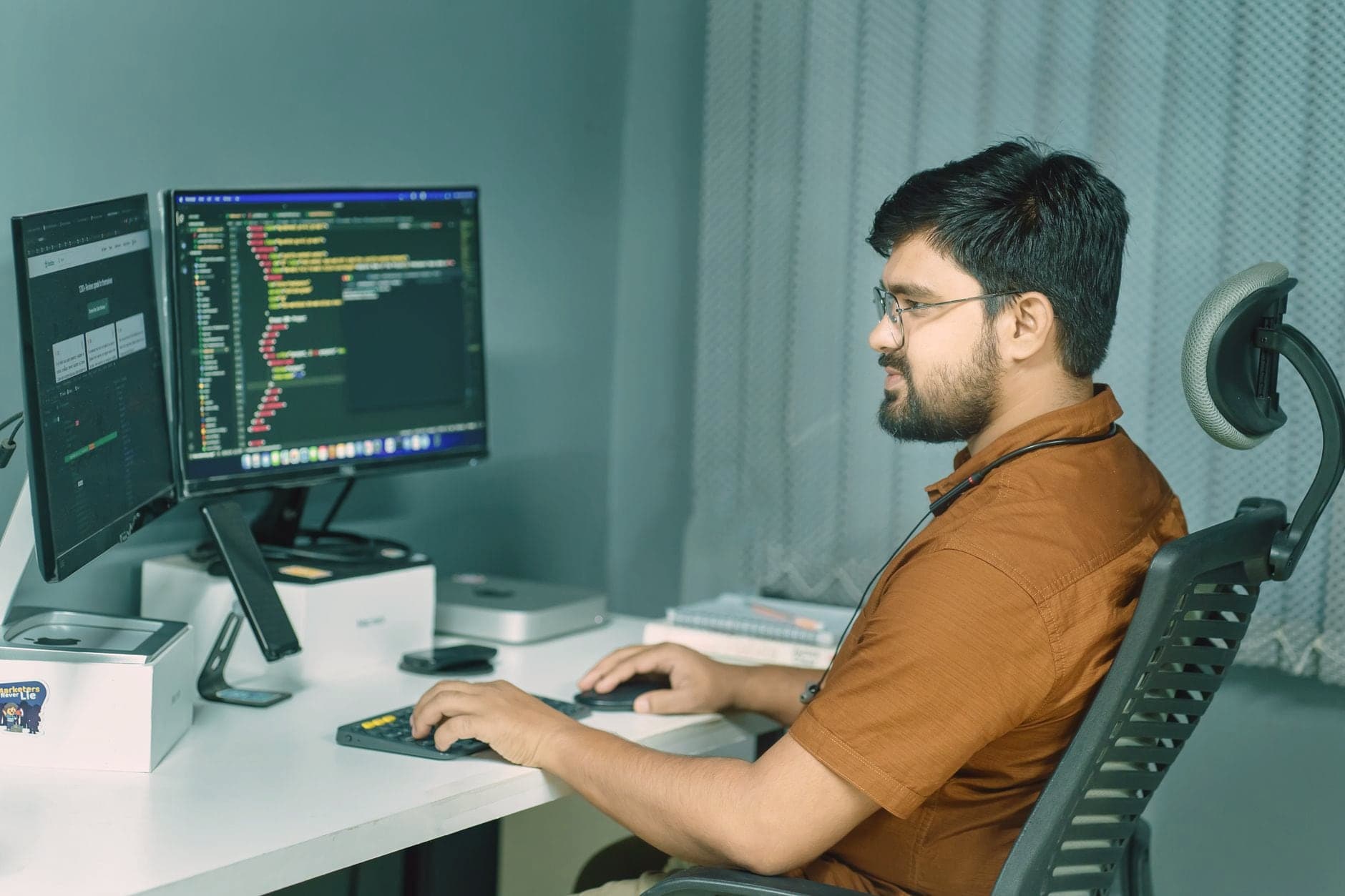 Developer with glasses writing code on two monitors at a white desk in an office with teal walls