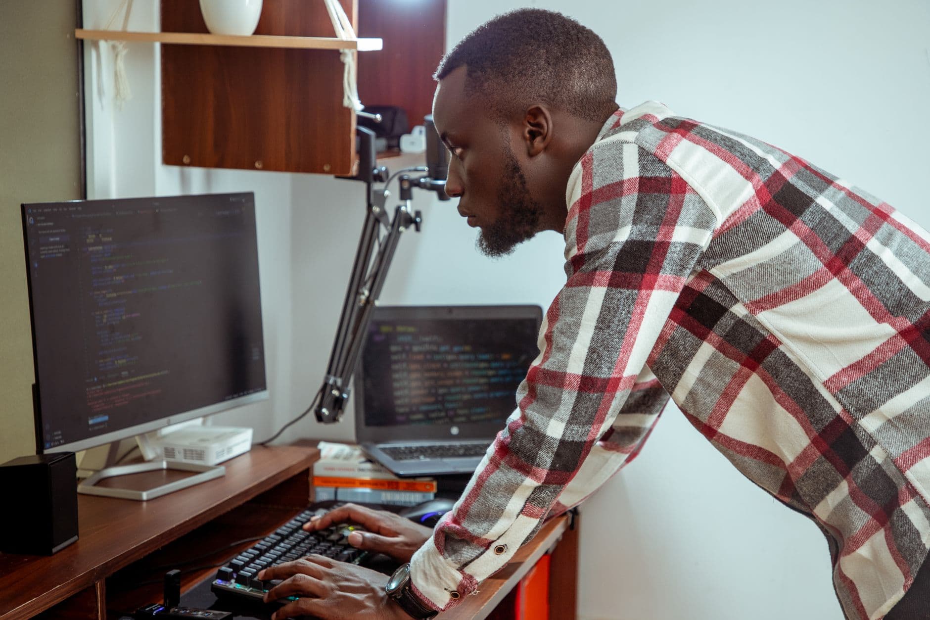 Developer leaning over his desk to type, with code on a monitor and an open laptop in front of him